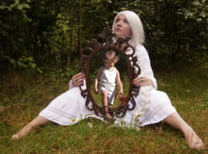 Jonge vrouw met wit haar en witte jurk zit in het gras. Ze houdt een spiegel vast waaruit een peuter stapt.